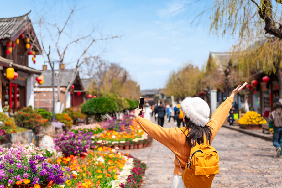 Mujer con mochila amarilla y sombrero contempla calle tradicional china con flores coloridas y linternas rojas.