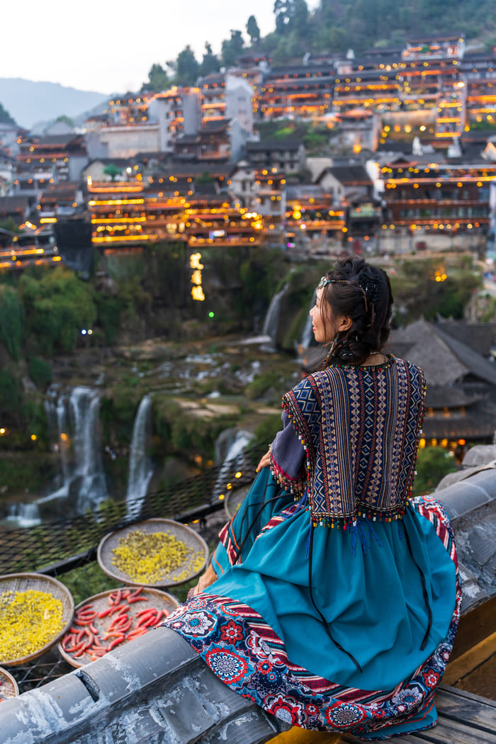 Mujer con vestimenta tradicional contempla el pueblo iluminado de Xijiang al atardecer, con arquitectura de madera en las montañas y cascadas en primer plano