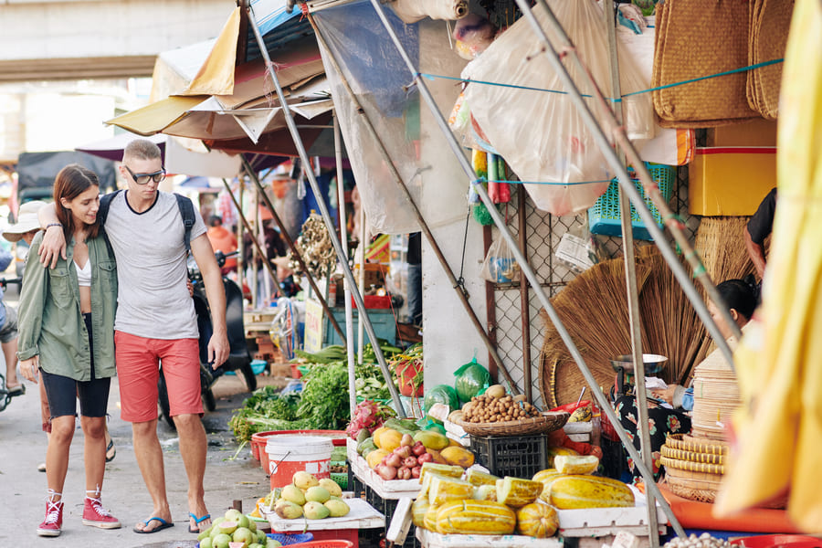 Pareja recorre mercado callejero asiático con puestos de frutas frescas y productos locales bajo toldos